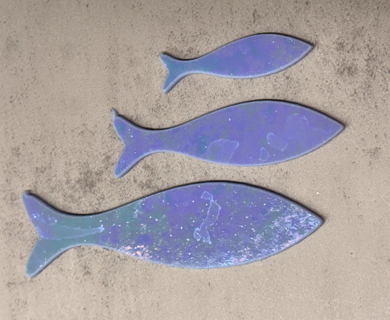 May include: Three iridescent blue fish cutouts of varying sizes are arranged on a textured, light brown surface. The fish have a shimmering, speckled appearance, with a gradient of blue and green hues. The fish tails are clearly defined.