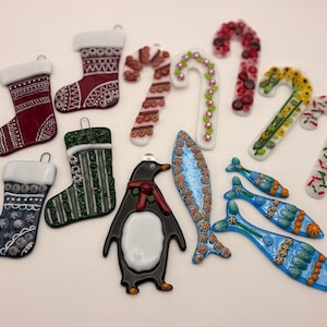 May include: A collection of glass Christmas ornaments in various shapes, including stockings, candy canes, a penguin, and fish. The ornaments feature detailed designs in red, green, blue, and white, with silver hanging loops.