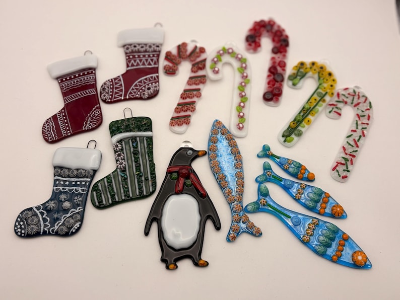 May include: A collection of glass Christmas ornaments in various shapes, including stockings, candy canes, a penguin, and fish. The ornaments feature detailed designs in red, green, blue, and white, with silver hanging loops.