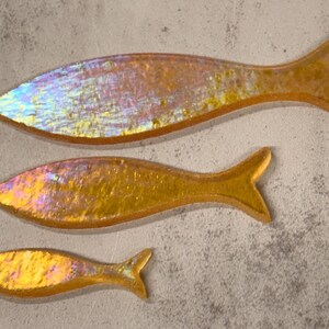 May include: Three iridescent fish-shaped decorative pieces in varying sizes. The fish have a golden-yellow hue with hints of pink and blue, and are arranged on a textured, light gray surface. The fish have a smooth, reflective surface.