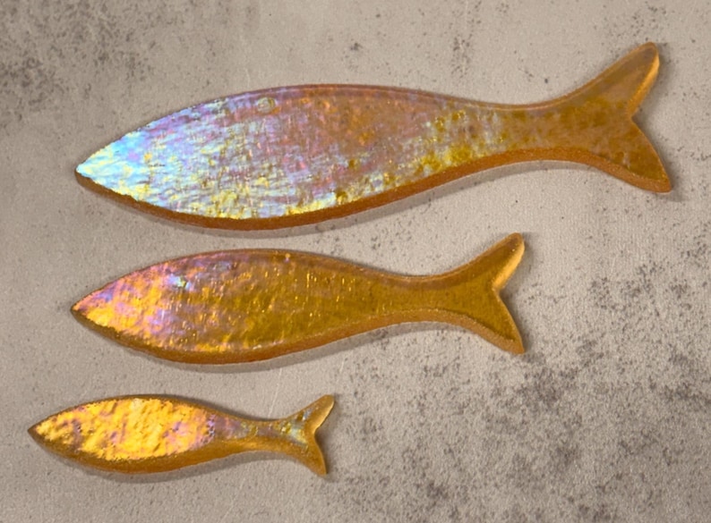 May include: Three iridescent fish-shaped decorative pieces in varying sizes. The fish have a golden-yellow hue with hints of pink and blue, and are arranged on a textured, light gray surface. The fish have a smooth, reflective surface.
