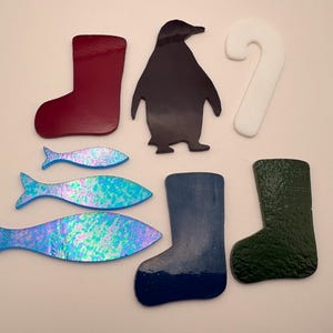 May include: A collection of colorful, die-cut shapes including a burgundy boot, a black penguin, a white candy cane, three iridescent blue fish, a blue boot, and a green textured boot. These shapes are likely for crafting or decoration.