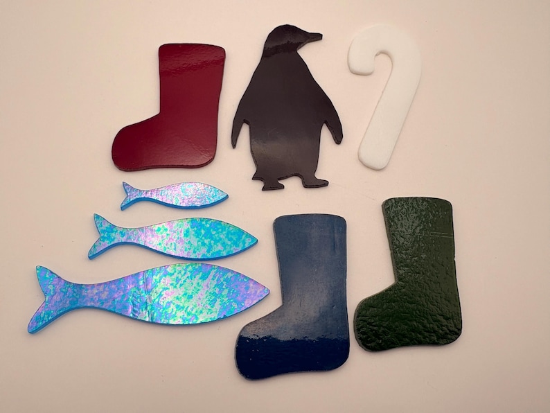 May include: A collection of colorful, die-cut shapes including a burgundy boot, a black penguin, a white candy cane, three iridescent blue fish, a blue boot, and a green textured boot. These shapes are likely for crafting or decoration.