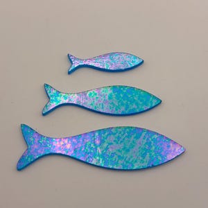 May include: Three iridescent fish cutouts in varying sizes. The fish have a blue outline and a shimmering surface with shades of blue, green, and purple. The fish are arranged vertically against a neutral background.