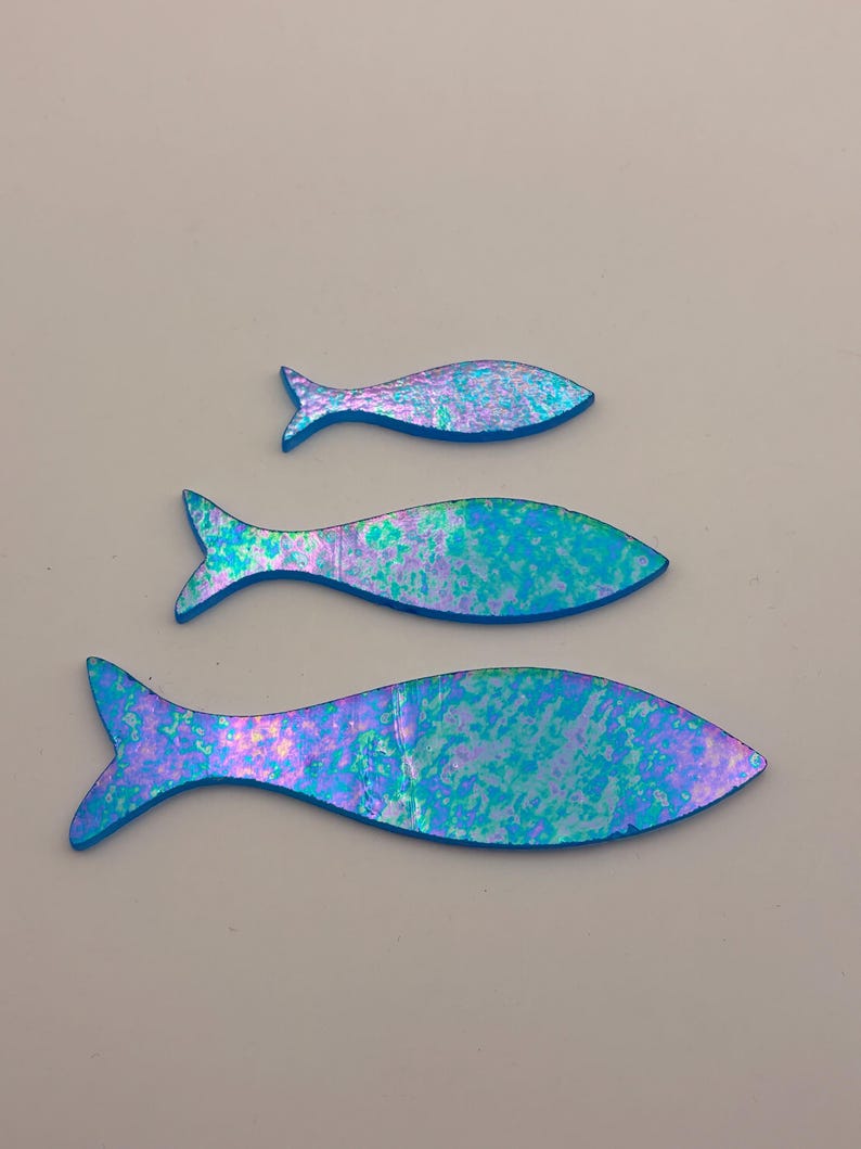 May include: Three iridescent fish cutouts in varying sizes. The fish have a blue outline and a shimmering surface with shades of blue, green, and purple. The fish are arranged vertically against a neutral background.