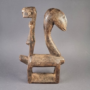 Puede incluir: Una escultura de madera de una persona montando una serpiente. La escultura está tallada en un estilo sencillo, con los brazos de la persona levantados y la cabeza de la serpiente girada hacia un lado. La escultura está pintada de un color marrón oscuro.