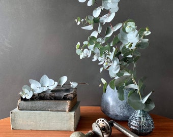 May include: A still life featuring a bouquet of eucalyptus branches in a gray ceramic vase, a stack of books, and a decorative door knob on a wooden table.