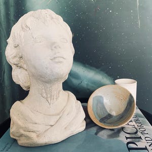 May include: A plaster bust of a young person with a serene expression. Beside it, a small ceramic bowl with blue and tan swirls and a white ceramic cup rest on a book titled "Picasso and Portraiture".