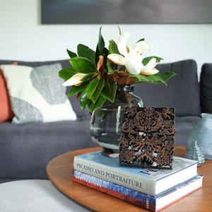 May include: A wooden block print with a detailed design sits on top of two books. The books are titled "Picasso and Portraiture" and "Destinations". A vase with white magnolia flowers and green leaves sits on a wooden table.