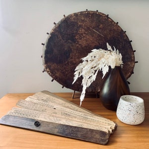 May include: A wooden fan with a brown handle and a paper fan with handwritten text and illustrations. The fan is on a wooden table with a brown vase with white dried flowers and a white speckled candle holder.