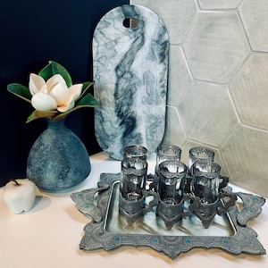 May include: A silver serving tray with ornate details and a mirrored surface. Six silver teacups with intricate designs are arranged on the tray. The tray is set on a white surface with a black and white marble chopping board in the background.