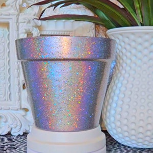 May include: A silver iridescent glitter planter pot with a white base. The pot is made of terracotta and has a smooth, shiny finish.