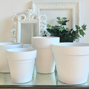 May include: A collection of five white ceramic plant pots of varying sizes. The pots have a matte finish and a simple, classic design. The background includes a white ornate picture frame and greenery.