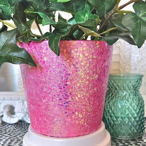 May include: A pink planter with a glittery finish. The planter has a white base and is filled with green leafy plants.