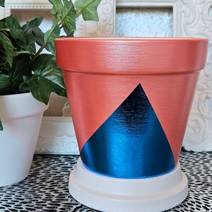 May include: A terracotta planter with a geometric design. The planter is painted with a blue triangle on a red background. The planter has a white base.