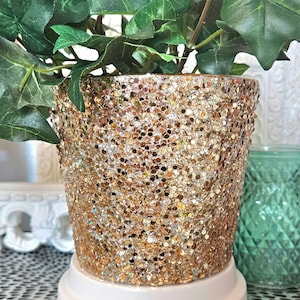 May include: A gold glitter planter with a green plant inside. The planter is sitting on a white pedestal.