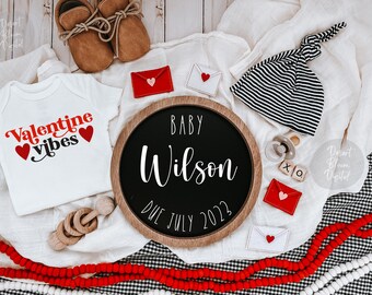 Può includere: Un body bianco con la scritta "Valentine vibes" in lettere rosse e nere e due cuori rossi. Un cartello rotondo di legno con la scritta "BABY Wilson DUE JULY 2022" in lettere bianche. Un cappello annodato a righe bianche e nere, un sonaglio di legno e buste rosse e bianche con cuori.