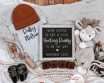 May include: A baby announcement with a letter board that reads "We're excited to say a little Hunting Buddy is on the way November 2025". The board is surrounded by a gray and white striped blanket, a brown knitted hat, a gray onesie with the text "Baby Wilson", a wooden rattle, a brown bottle, a pair of camouflage booties, a wooden bead garland, a crocheted deer toy, and wooden blocks that spell "BABY".