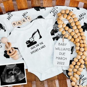 Puede incluir: Un body blanco con un gráfico de excavadora negro y el texto "Here for the ditches & hoes". Una etiqueta blanca con el texto "Baby Williams Due March 2022" cuelga de una hebra de cuentas de madera. Una manta de bebé en blanco y negro con un patrón geométrico está en el fondo.