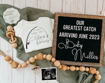 Op de afbeelding: Een witte romper met een afbeelding van een hengel en de tekst "Ik ben het bewijs dat papa niet altijd aan het vissen is". Op de achtergrond is een letterbord met de tekst "Onze grootste vangst arriveert in juni 2023 Baby Miller".