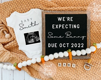 Peut inclure: Une grenouillère blanche avec le texte "Baby Smith" et une image d'échographie en noir et blanc. Un tableau noir avec le texte "We're expecting some bunny due Oct 2022". Une guirlande de pompons blancs avec des carottes orange.