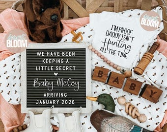 May include: A baby announcement with a white onesie printed with "I'M PROOF DADDY ISN'T HUNTING ALL THE TIME." Includes a letter board with the text "WE HAVE BEEN KEEPING A LITTLE SECRET Baby McCoy ARRIVING JANUARY 2026." Also includes baby boots, wooden blocks, and a stuffed dog.