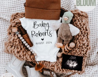 May include: A baby announcement featuring a white onesie with "Baby Roberts" and "August 2026" printed on it. The announcement includes a brown beanie, a plush duck, a pacifier, a pair of white boots, and an ultrasound image, all arranged on a textured blanket.