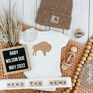 May include: A white onesie with a brown buffalo silhouette, a wooden letter board that reads "BABY WILSON DUE MAY 2022", a brown knitted blanket, a wooden rattle, a wooden bead garland, and a brown knitted beanie.
