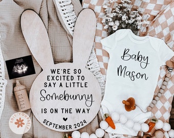 May include: A wooden bunny-shaped sign with the text "We're so excited to say a little Somebunny is on the way September 2024". A white onesie with the text "Baby Mason" is next to the sign. The sign and onesie are surrounded by white and brown decorative items.