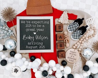 May include: A red baby onesie with a black letter board that reads "We're expecting 2025 to be a great year Baby Wilson arriving August 2025". There are wooden blocks that spell "BABY" and a wooden ornament that reads "Poppin' Bottles of a different kind".