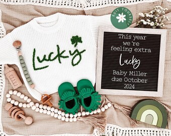 May include: A white knitted baby sweater with the word "Lucky" embroidered in green yarn. The sweater is surrounded by a variety of baby accessories, including a wooden rattle, a green and white beaded necklace, a pair of green baby shoes, and a letter board that reads "This year we're feeling extra Lucky Baby Miller due October 2024".