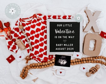 Può includere: Una lavagna nera con il testo "Our little Valentine is on the way. Baby Miller August 2024." Un body bianco con cuori rossi, un sonaglio di legno, una collana di perline di legno e una piccola immagine di ecografia sono disposti intorno alla lavagna.