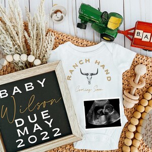 Könnte beinhalten: Ein weißer Strampler mit dem Text "RANCH HAND Coming Soon" und einem Totenkopf- und Knochen-Design. Ein Schwarzweiß-Ultraschallbild befindet sich auf dem Strampler. Ein Holzschild mit dem Text "BABY Wilson DUE MAY 2022" ist im Bild.