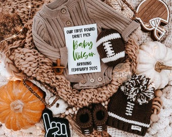May include: A baby announcement featuring a brown knit onesie, football-themed accessories, and a sign that reads "OUR FIRST ROUND DRAFT PICK Baby Wilson ARRIVING FEBRUARY 2026." The arrangement includes a football, a beanie, and booties.