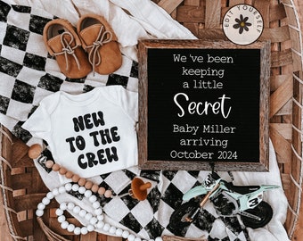 May include: A black and white checkered blanket with a white onesie that says "New to the Crew" in black. A small wooden sign with the text "We've been keeping a little Secret Baby Miller arriving October 2024" is on the blanket. There are brown baby shoes, a wooden bead necklace, and a small toy motorcycle on the blanket.