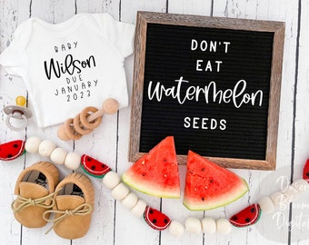 May include: A white onesie with the text "Baby Wilson Due January 2023" on it. A wooden rattle, a pacifier, and a string of white beads with watermelon slices are also in the image. A black letter board with the text "Don't Eat Watermelon Seeds" is in the background.