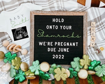 May include: A black letter board with the text "Hold onto your Shamrocks We're Pregnant Due June 2022". A white onesie with the text "Baby Smith" and an ultrasound image is in the background. Green shamrock decorations surround the letter board.