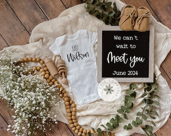 May include: A white onesie with the text "BABY Wilson" on it, a wooden rattle, a wooden bottle, a string of wooden beads, a letter board with the text "We can't wait to meet you June 2024", a bouquet of white flowers, and a pair of brown baby shoes.