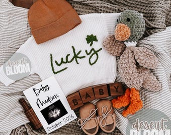 May include: A baby announcement featuring a white sweater with "Lucky" and a shamrock, a brown beanie, a crochet duck, baby shoes, wooden blocks spelling "BABY", and a sonogram photo. The announcement is for October 2026.
