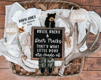 May include: A rustic baby announcement featuring a white onesie with "BABY JONES DUE MARCH 2026" printed on it, a black teddy bear, and a sign that reads "RIFLES, RACKS & Deer Tracks THAT'S WHAT LITTLE BOYS are made of".
