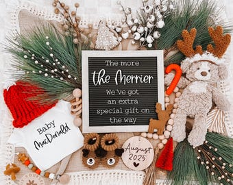 May include: A Christmas-themed baby announcement with a letter board that reads "The more the merrier. We've got an extra special gift on the way."  The board is surrounded by a red and white Santa hat, a white onesie with the text "Baby MacDonald", a pair of brown baby booties, a wooden reindeer ornament, a wooden sign with the text "August 2025", and a brown teddy bear wearing a reindeer headband.
