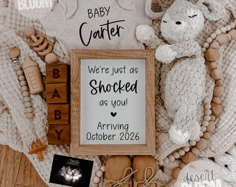 May include: A baby announcement featuring a gray onesie with "BABY Carter" printed on it, a wooden framed sign that reads "We're just as shocked as you! Arriving October 2026", and a plush deer toy. Also included are baby shoes, blocks that spell "BABY", and a pacifier.