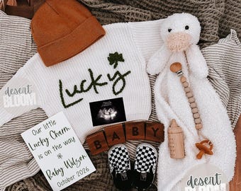 May include: A baby announcement featuring a white sweater with "Lucky" embroidered in green, a brown beanie, and checkered baby shoes. Includes a sonogram image, wooden blocks spelling "BABY", and a card announcing "Baby Wilson" for October 2026.