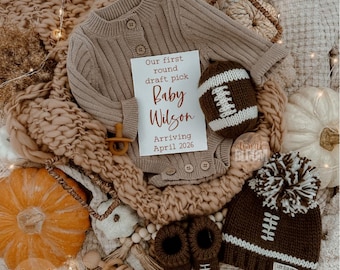 May include: A neutral-toned baby announcement featuring a knit football, hat, and booties. A onesie and pacifier are also present. A card reads "Our first round draft pick Baby Wilson Arriving April 2026."