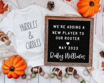 May include: A white onesie with the text "HUDDLES & CUDDLES" on it. A wooden letter board with the text "WE'RE ADDING A NEW PLAYER TO OUR ROSTER MAY 2023 Baby Williams" is next to the onesie. The letter board is surrounded by fall-themed decorations, including pumpkins, leaves, and pine cones.