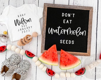May include: A white onesie with the text "Baby Wilson Due January 2023" on it, a letter board with the text "Don't Eat Watermelon Seeds", a pair of black and white polka dot baby shoes, a wooden rattle, a pacifier, and a watermelon garland.