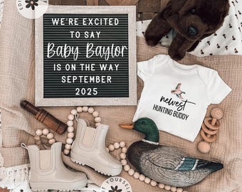 Puede incluir: Un body blanco con el texto "nuevo compañero de caza" y una pequeña imagen de un pato. Un tablero de letras con el texto "Estamos emocionados de decir que Baby Baylor está en camino para septiembre de 2025". Un señuelo de pato de madera, un par de botas de lluvia beige, un sonajero de madera y un perro de peluche marrón.