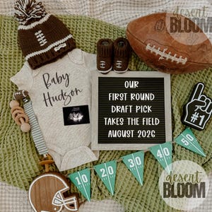May include: A baby announcement with a football theme. Features a brown and white knitted hat, matching booties, and a onesie with the name "Baby Hudson." Includes a football, a letter board, and a banner with yard markers.