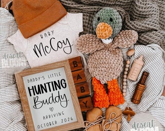 May include: A collection of baby items, including a tan beanie, a onesie with "Baby McCoy" printed on it, and a framed sign that reads "Daddy's Little Hunting Buddy Arriving October 2026." Also included is a crochet duck, baby shoes, and a wooden baby bottle.