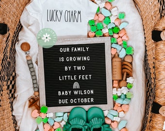 May include: A black letter board with the text "Our family is growing by two little feet Baby Wilson Due October". Green baby booties, a white onesie with the text "Lucky Charm", and a wooden rattle are on a woven basket with green, pink, and white candy.